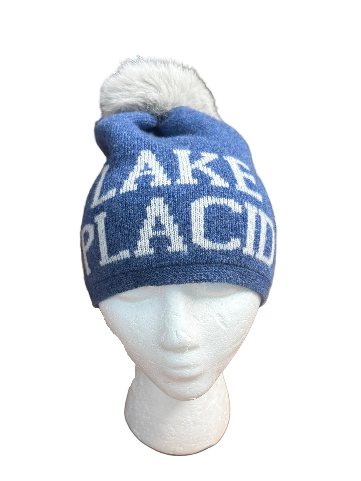A blue knit beanie with a white pom-pom and white lettering that reads 'LAKE PLACID' displayed on a white mannequin head, set on a brown table with a store interior in the background.