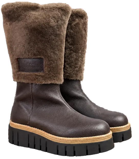 A pair of dark brown leather ankle boots with a thick brown shearling collar and a chunky black lug sole with a tan midsole, shown on a plain white background.