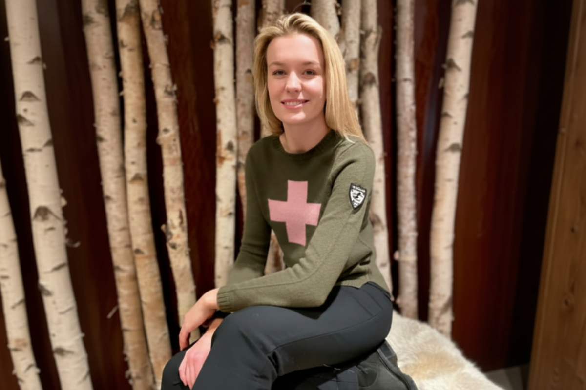 blonde woman sitting and wearing an olive green sweater with a pale pink cross on chest and a patch showing a skier on the upper arm of the sleeve.