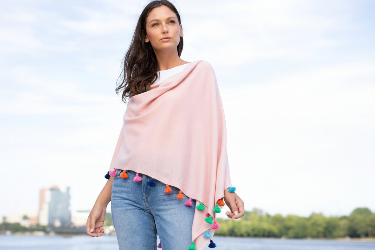 A woman wearing a navy blue cotton cashmere festive tassel trim topper with multi colored tassels, paired with jeans.